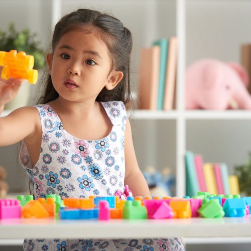 Little girl playing with plastick blocks at table in nursery room