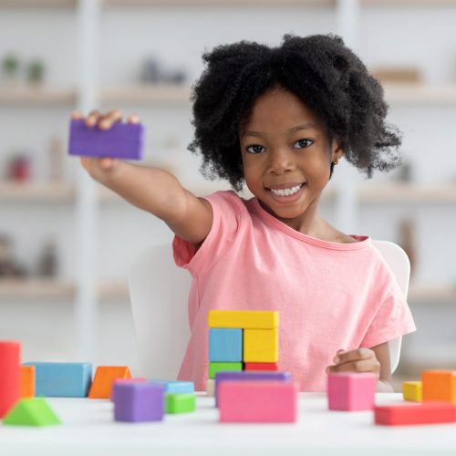 Cute little black girl preschooler with bushy hair showing purple wooden block at camera, happy child having fun at kindergarten, playing with bricks, making construction, kids education concept